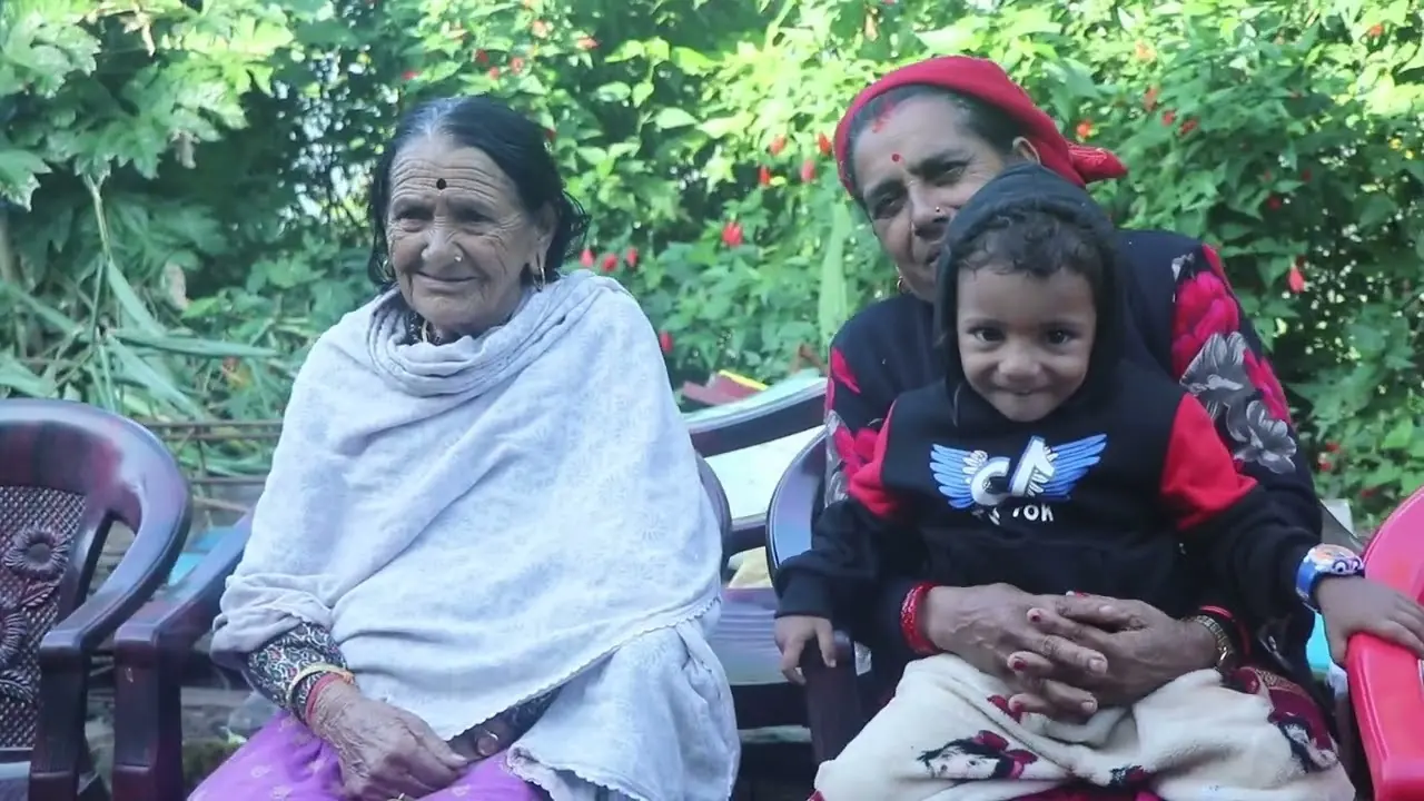 An elderly woman in a white shawl sits beside a younger relative holding a small child, in a garden during a chaurasi puja