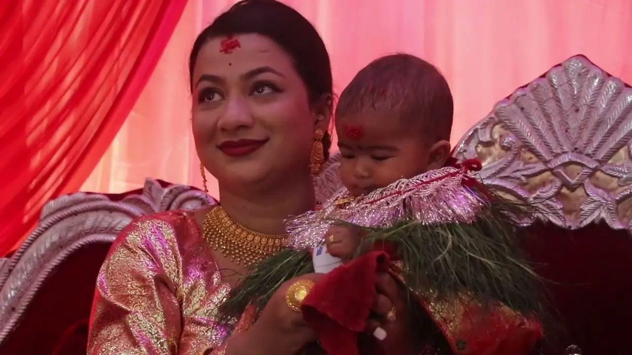 A smiling woman in red with tika on her forehead holds a baby during a pasni ceremony in Walling